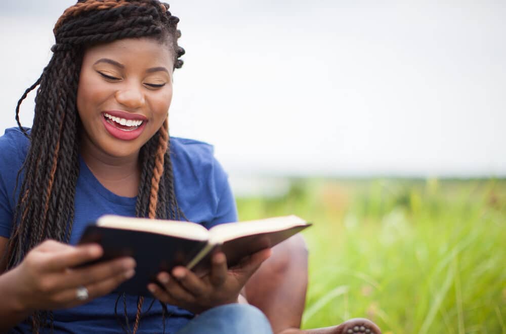 An African-American woman smiling while reading the Bible outside in a field.
